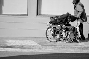 A person in a hooded jacket is seated in a wheelchair on a paved sidewalk, with another individual standing nearby holding crutches. The scene is in black and white, with a plain building facade in the background.