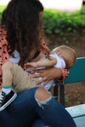 A woman seated on a green bench is breastfeeding a baby. She is wearing a pink jacket with black polka dots and jeans with a tear at the knee. The baby is dressed in a white shirt and beige pants, with one shoe visible. The background includes greenery and blurred natural elements, suggesting an outdoor setting.