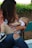 A woman seated on a green bench is breastfeeding a baby. She is wearing a pink jacket with black polka dots and jeans with a tear at the knee. The baby is dressed in a white shirt and beige pants, with one shoe visible. The background includes greenery and blurred natural elements, suggesting an outdoor setting.