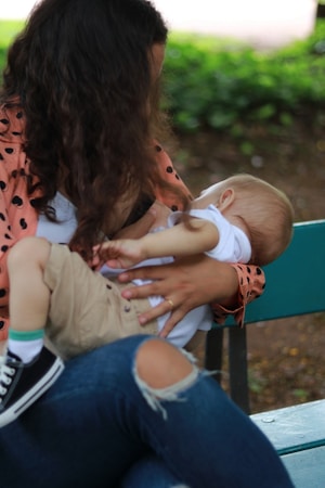 A woman seated on a green bench is breastfeeding a baby. She is wearing a pink jacket with black polka dots and jeans with a tear at the knee. The baby is dressed in a white shirt and beige pants, with one shoe visible. The background includes greenery and blurred natural elements, suggesting an outdoor setting.