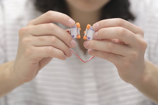 Hands are holding electronic components connected with a red wire. The components have white bases with orange connectors, and the background is softly blurred.
