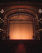An ornate theater interior featuring an elaborately decorated proscenium arch with intricate artwork and carvings. The stage is adorned with a large, golden curtain that extends across its width. Rows of red velvet seats line the seating area, and the ambiance is dimly lit by elegant chandeliers and lights along the walls.