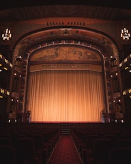 An ornate theater interior featuring an elaborately decorated proscenium arch with intricate artwork and carvings. The stage is adorned with a large, golden curtain that extends across its width. Rows of red velvet seats line the seating area, and the ambiance is dimly lit by elegant chandeliers and lights along the walls.