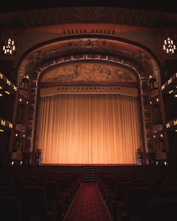An ornate theater interior featuring an elaborately decorated proscenium arch with intricate artwork and carvings. The stage is adorned with a large, golden curtain that extends across its width. Rows of red velvet seats line the seating area, and the ambiance is dimly lit by elegant chandeliers and lights along the walls.