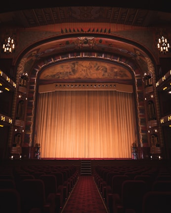 An ornate theater interior featuring an elaborately decorated proscenium arch with intricate artwork and carvings. The stage is adorned with a large, golden curtain that extends across its width. Rows of red velvet seats line the seating area, and the ambiance is dimly lit by elegant chandeliers and lights along the walls.
