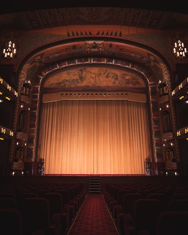 An ornate theater interior featuring an elaborately decorated proscenium arch with intricate artwork and carvings. The stage is adorned with a large, golden curtain that extends across its width. Rows of red velvet seats line the seating area, and the ambiance is dimly lit by elegant chandeliers and lights along the walls.