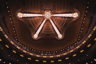 A grand, intricately designed ceiling featuring a central chandelier with ornate patterns and geometric lighting elements. The structure is surrounded by tiers of decorative lighting that add a warm, inviting glow.