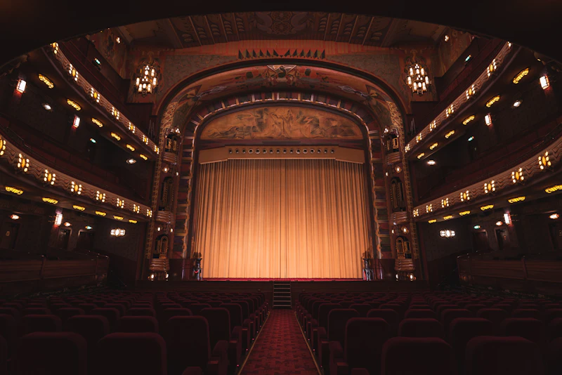 An ornate theater with rows of red seats facing a lit stage and closed gold curtain