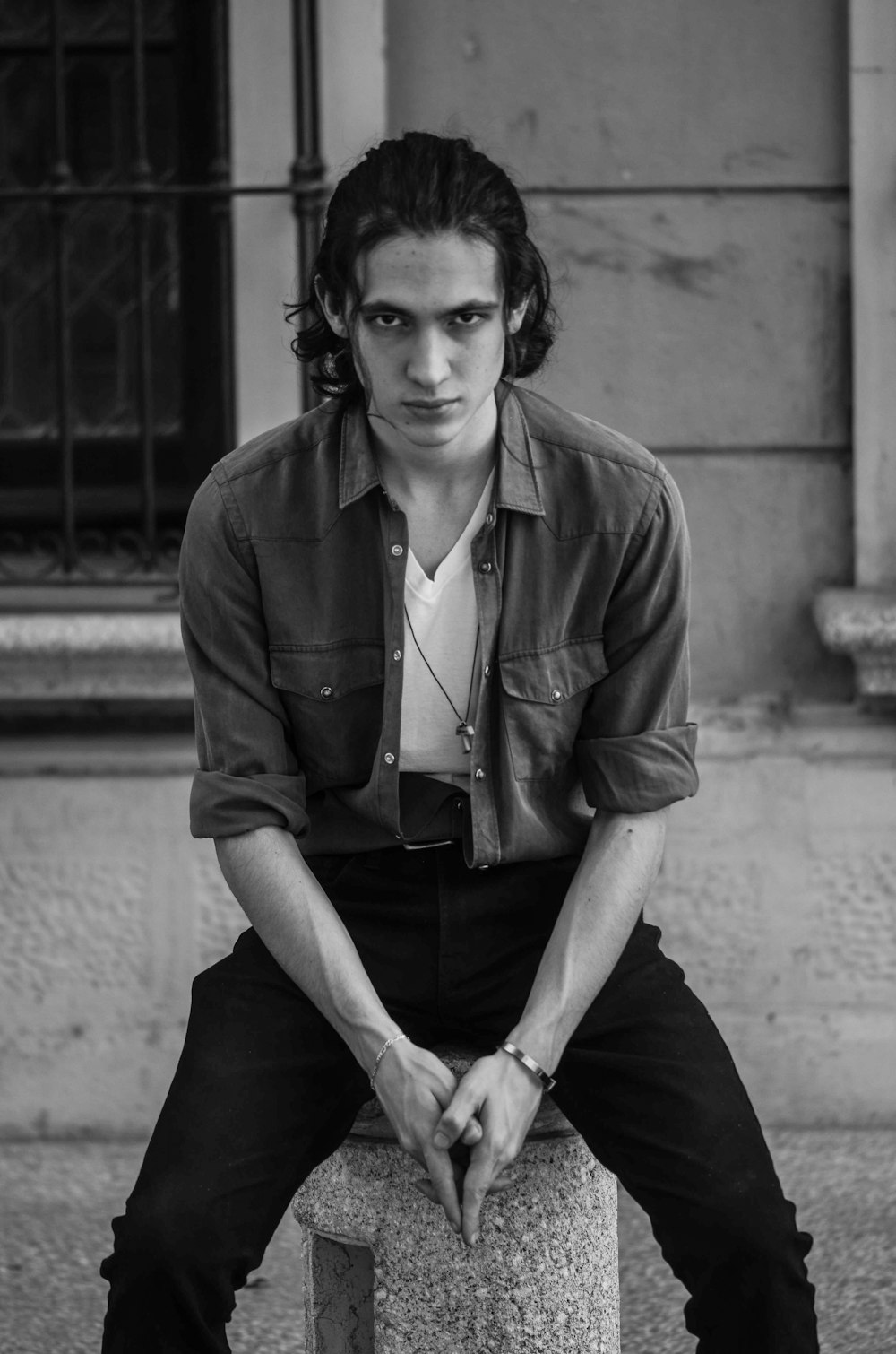 grayscale photo of man in button up shirt and black pants sitting on concrete wall