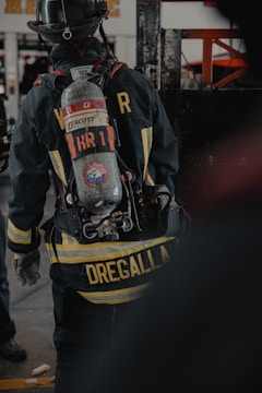 A firefighter is seen from the back, wearing full gear including a helmet and an oxygen tank. The jacket displays a prominent yellow striping and a name tag, with equipment attached to the waist belt. The environment appears to be indoors, possibly a fire station or training facility.