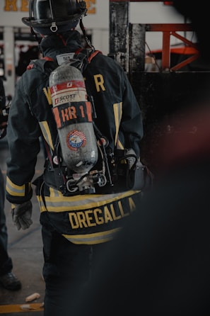 A firefighter is seen from the back, wearing full gear including a helmet and an oxygen tank. The jacket displays a prominent yellow striping and a name tag, with equipment attached to the waist belt. The environment appears to be indoors, possibly a fire station or training facility.