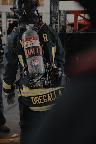 A firefighter is seen from the back, wearing full gear including a helmet and an oxygen tank. The jacket displays a prominent yellow striping and a name tag, with equipment attached to the waist belt. The environment appears to be indoors, possibly a fire station or training facility.