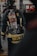 A firefighter is seen from the back, wearing full gear including a helmet and an oxygen tank. The jacket displays a prominent yellow striping and a name tag, with equipment attached to the waist belt. The environment appears to be indoors, possibly a fire station or training facility.