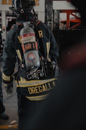 A firefighter is seen from the back, wearing full gear including a helmet and an oxygen tank. The jacket displays a prominent yellow striping and a name tag, with equipment attached to the waist belt. The environment appears to be indoors, possibly a fire station or training facility.