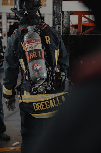A firefighter is seen from the back, wearing full gear including a helmet and an oxygen tank. The jacket displays a prominent yellow striping and a name tag, with equipment attached to the waist belt. The environment appears to be indoors, possibly a fire station or training facility.