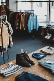 An interior of a clothing store with racks of various types of clothes, including jackets and shirts, hangs in the background. A wooden table in the foreground displays neatly arranged clothing items such as a pair of brown boots, a beanie hat, folded sweaters, and jeans.