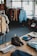 An interior of a clothing store with racks of various types of clothes, including jackets and shirts, hangs in the background. A wooden table in the foreground displays neatly arranged clothing items such as a pair of brown boots, a beanie hat, folded sweaters, and jeans.