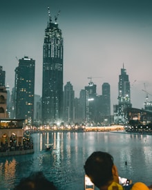 A cityscape featuring tall skyscrapers with varying designs dominates the backdrop, set against a slightly overcast sky. The skyline is illuminated by numerous lights, reflecting on the water body in the foreground. A small boat can be seen navigating the water, with people gathered along the waterfront. The scene is viewed from behind a person taking a photo with a smartphone.