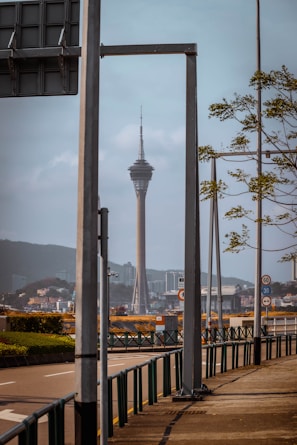 A tall telecommunications tower rises in the distance, framed by metal structures and signs. The foreground features a roadway lined with metal railings and sparse foliage. The cityscape is visible beneath a calm sky, with distant buildings and hills.