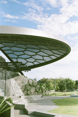 A modern architectural structure with a circular, honeycomb-patterned metal canopy. It is partially overhanging a textured wall with geometric designs, adorned with patches of greenery. The scene is set in a well-maintained, open park area with trees and a clear blue sky in the background.