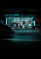 A small, brightly lit newsstand is surrounded by darkness on a city street. The stand is filled with a variety of magazines and newspapers. A person stands inside among the publications, while a car is parked nearby on the dimly lit street.