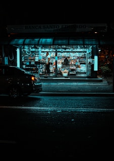 A small, brightly lit newsstand is surrounded by darkness on a city street. The stand is filled with a variety of magazines and newspapers. A person stands inside among the publications, while a car is parked nearby on the dimly lit street.