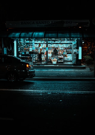 A small, brightly lit newsstand is surrounded by darkness on a city street. The stand is filled with a variety of magazines and newspapers. A person stands inside among the publications, while a car is parked nearby on the dimly lit street.