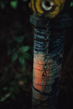 A close-up view of an old, weathered metal pipe with various types of faded text and graphic symbols. The pipe is vertically oriented and has a chain partially wrapped around it. The paint is chipped and rusted with a noticeable area of orange and red hues. The dim lighting and greenery partially visible in the soft-focus background add a mysterious or neglected atmosphere.