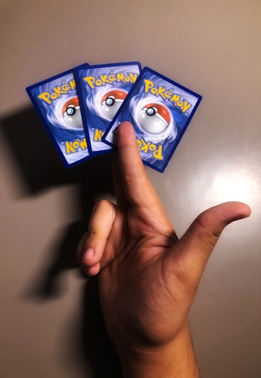 A hand holding three Pok&eacute;mon cards in a fan-like arrangement against a plain background. The cards are aligned with the Pok&eacute;mon logo and pok&eacute;ball design facing upwards.