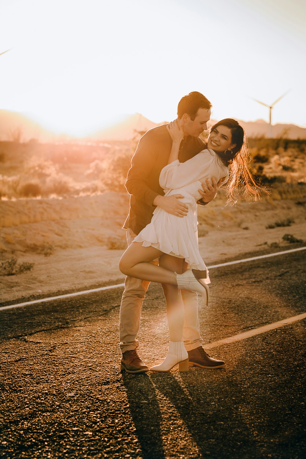 man in white tank top and white pants kissing woman in white dress on road during