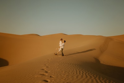 A couple embracing on expansive, golden sand dunes under a clear blue sky. The scene conveys a sense of romance and adventure, with footprints leading to their spot and long shadows cast by the setting sun.