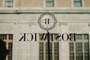 A glass window reflects an outdoor scene, featuring the word 'BOSTWICK' with the letter 'B' encased in a geometric design. The reflection includes a building with brick and concrete surfaces and several windows.