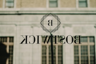 A glass window reflects an outdoor scene, featuring the word 'BOSTWICK' with the letter 'B' encased in a geometric design. The reflection includes a building with brick and concrete surfaces and several windows.