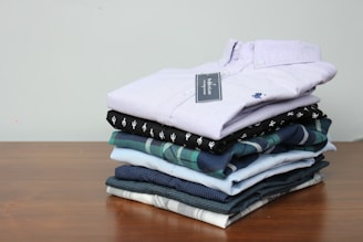 A neatly stacked pile of folded shirts is arranged on a wooden surface. The shirts vary in color and pattern, with one having a checked design and others in solid colors. The top shirt is a light lavender shade with a visible tag attached.