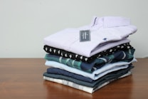 A neatly stacked pile of folded shirts is arranged on a wooden surface. The shirts vary in color and pattern, with one having a checked design and others in solid colors. The top shirt is a light lavender shade with a visible tag attached.