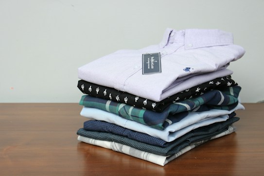 A neatly stacked pile of folded shirts is arranged on a wooden surface. The shirts vary in color and pattern, with one having a checked design and others in solid colors. The top shirt is a light lavender shade with a visible tag attached.