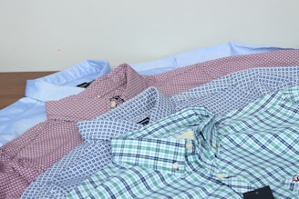 Several neatly folded button-up shirts in various patterns and colors are laid out on a surface. The shirts feature plaid, polka dot, and geometric designs in hues like blue, red, white, and green. A light blue shirt can be seen at the back, while the darker and more colorful patterns are in the front.