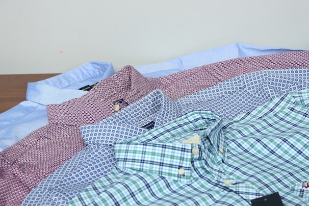 Several neatly folded button-up shirts in various patterns and colors are laid out on a surface. The shirts feature plaid, polka dot, and geometric designs in hues like blue, red, white, and green. A light blue shirt can be seen at the back, while the darker and more colorful patterns are in the front.