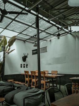 A modern, industrial-style café interior with wooden tables and chairs arranged neatly. The walls are adorned with a deer head silhouette and minimalist decorations. The ceiling is made of corrugated metal sheets, and there are large plants adding a touch of greenery. The overall ambiance is calm and cozy.