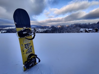 A snowboard is standing upright on a pristine snow-covered field. In the distance, there are snow-covered trees and a few traditional alpine houses. The sky is partly cloudy with patches of blue peeking through, and mountains are visible on the horizon.