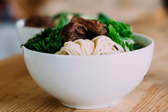 A white bowl filled with noodles, green leafy vegetables, and chunks of braised meat, placed on a wooden surface. The dish includes fresh ingredients that are well-prepared and appetizing.