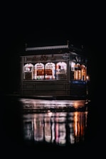 An illuminated food kiosk is situated in the darkness, with colorful snacks and drinks visible through its windows. The kiosk's lights reflect on the wet surface below, creating a vibrant mirrored effect.
