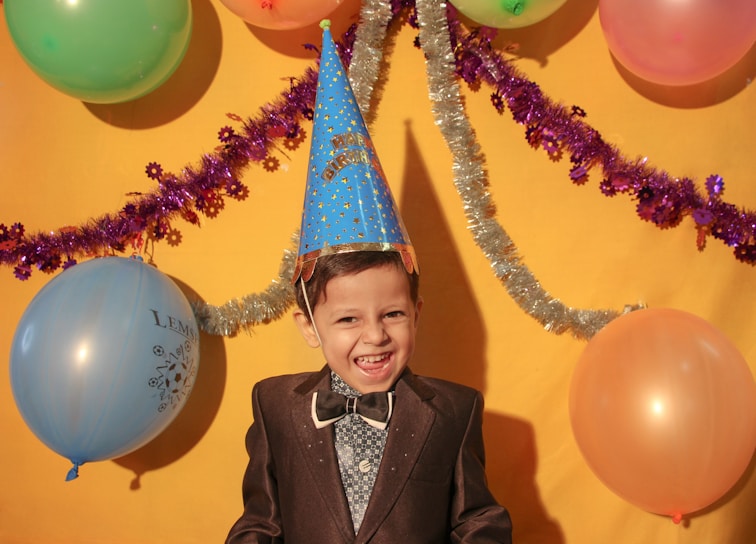 A young child wearing a party hat and dressed in a suit stands in front of a bright yellow backdrop. The scene is decorated with colorful balloons and shiny streamers, creating a festive atmosphere. The child is smiling broadly, adding a sense of joy to the celebration.