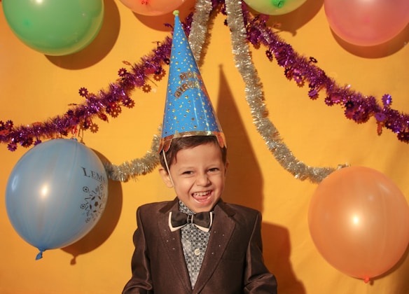 A young child wearing a party hat and dressed in a suit stands in front of a bright yellow backdrop. The scene is decorated with colorful balloons and shiny streamers, creating a festive atmosphere. The child is smiling broadly, adding a sense of joy to the celebration.