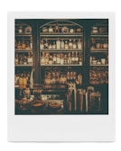 Dimly lit bar shelves stocked with various bottles of alcohol and glassware. Metal cocktail shakers are visible on the counter, along with some small bowls and containers.