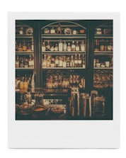 Dimly lit bar shelves stocked with various bottles of alcohol and glassware. Metal cocktail shakers are visible on the counter, along with some small bowls and containers.
