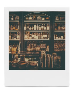 Dimly lit bar shelves stocked with various bottles of alcohol and glassware. Metal cocktail shakers are visible on the counter, along with some small bowls and containers.