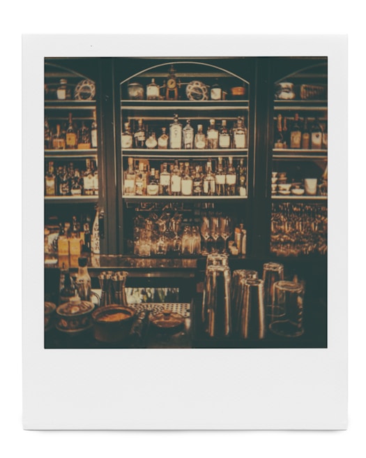 Dimly lit bar shelves stocked with various bottles of alcohol and glassware. Metal cocktail shakers are visible on the counter, along with some small bowls and containers.