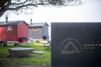 A scenic setting with two small, house-like structures on wheels, resembling compact mobile homes or trailers. One is painted red and the other gray, both equipped with chimneys. A tree partially frames the scene from the left, casting shadows on the grass below. A prominent signboard labeled 'Nagasaki Beach Resort' stands on the right, featuring a minimalist emblem.