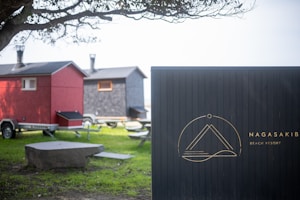 A scenic setting with two small, house-like structures on wheels, resembling compact mobile homes or trailers. One is painted red and the other gray, both equipped with chimneys. A tree partially frames the scene from the left, casting shadows on the grass below. A prominent signboard labeled 'Nagasaki Beach Resort' stands on the right, featuring a minimalist emblem.